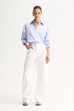 Model wearing the Light Blue Elka Collective Carter Shirt with white wide-leg jeans and black thong sandals, showing the classic shirt collar and front monogram detail, full-length front view