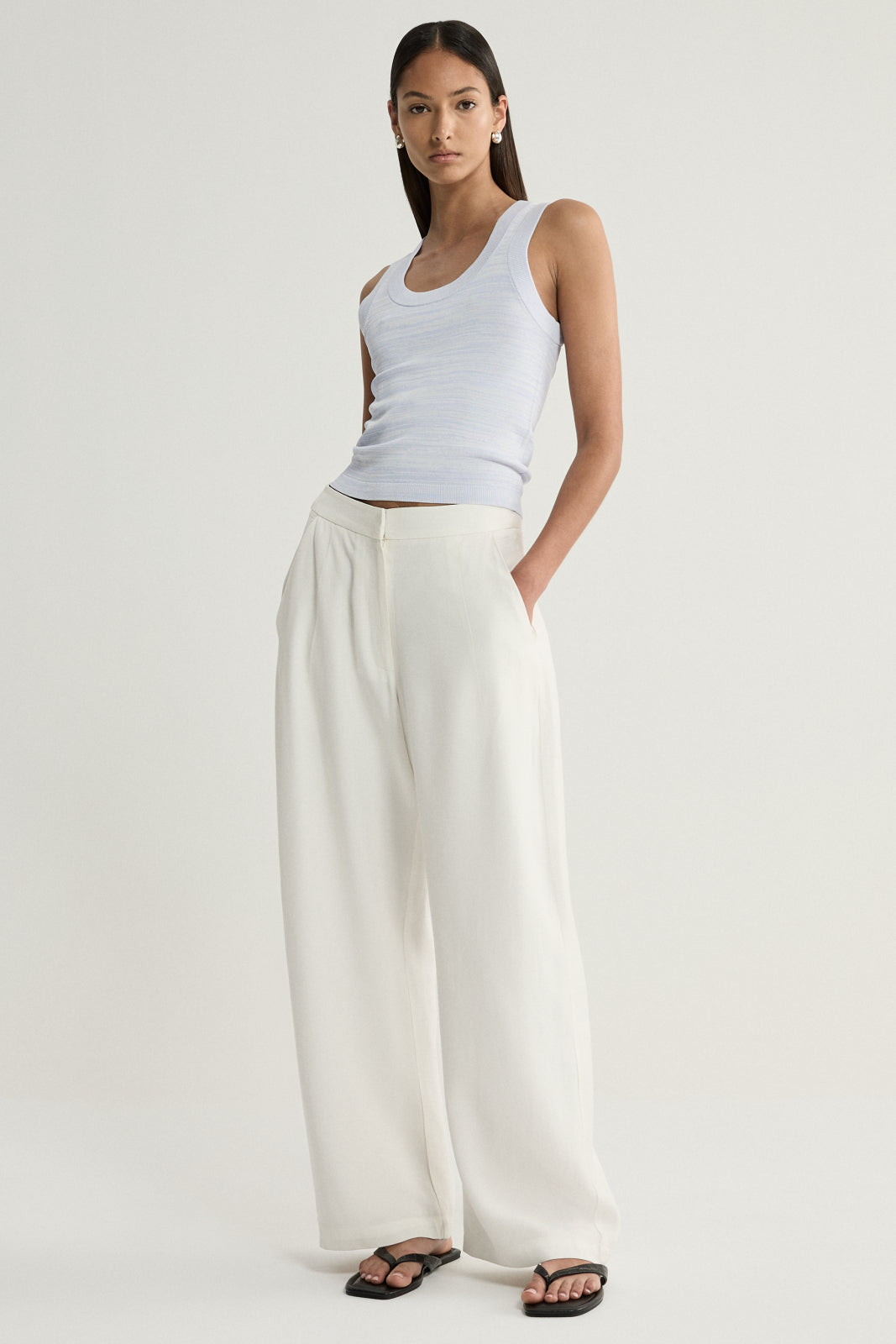 A model wearing a pale blue marled sleeveless knit top with a scoop neckline, paired with cream wide-leg trousers and black flat sandals in a full-length front view.