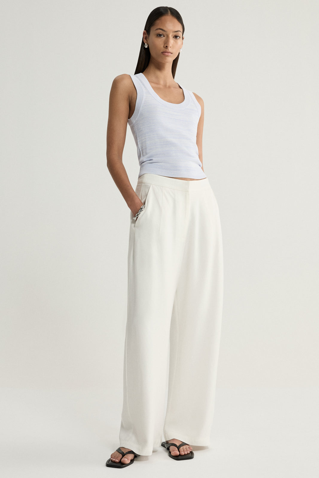 A model wearing a pale blue marled sleeveless top with a rounded scoop neckline and cream wide-leg trousers, standing with one hand in a pocket and a silver bracelet at the wrist.