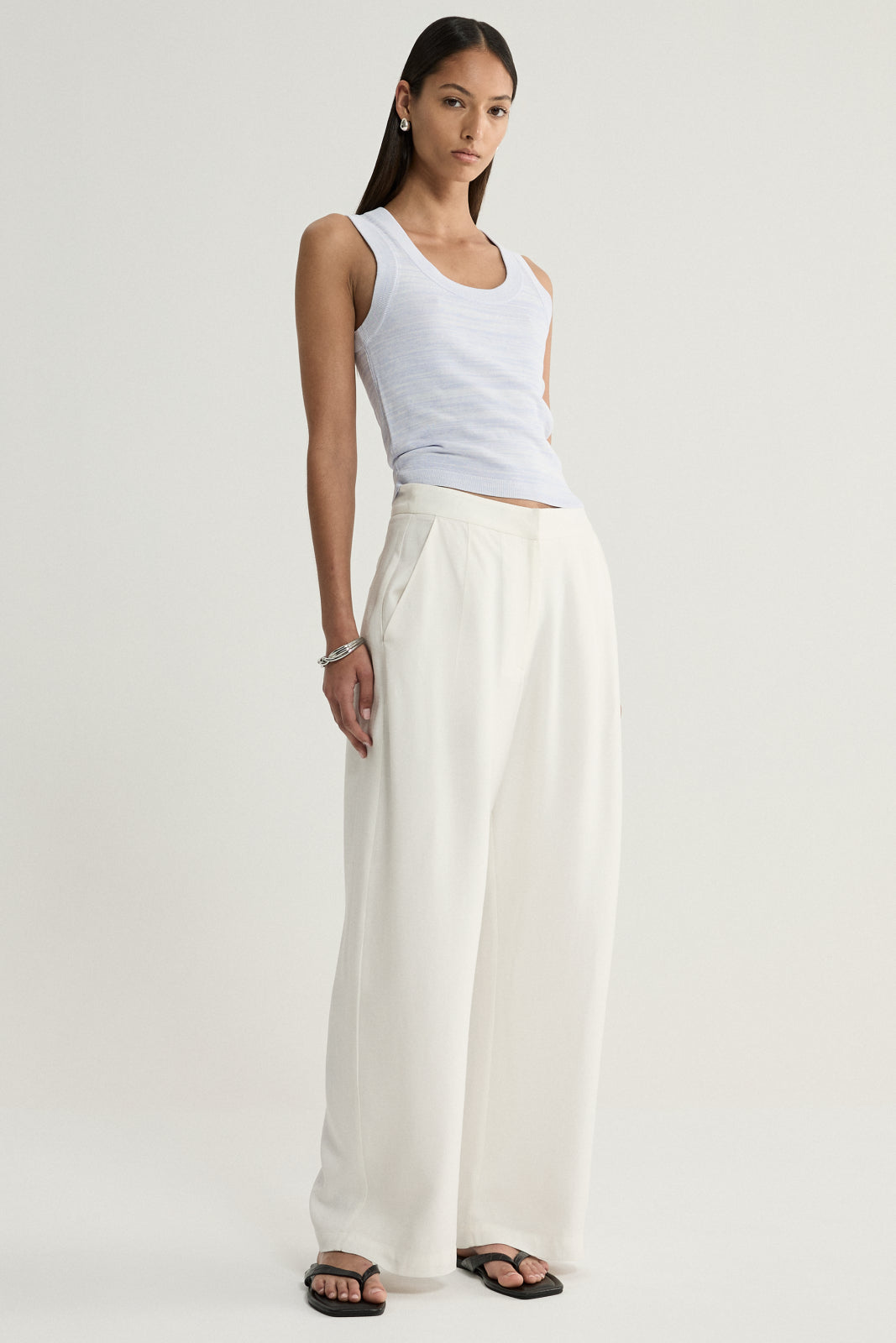 A model wearing a pale blue marled knit tank with a scoop neckline and fitted shape, paired with cream wide-leg trousers and black sandals in a relaxed standing pose.