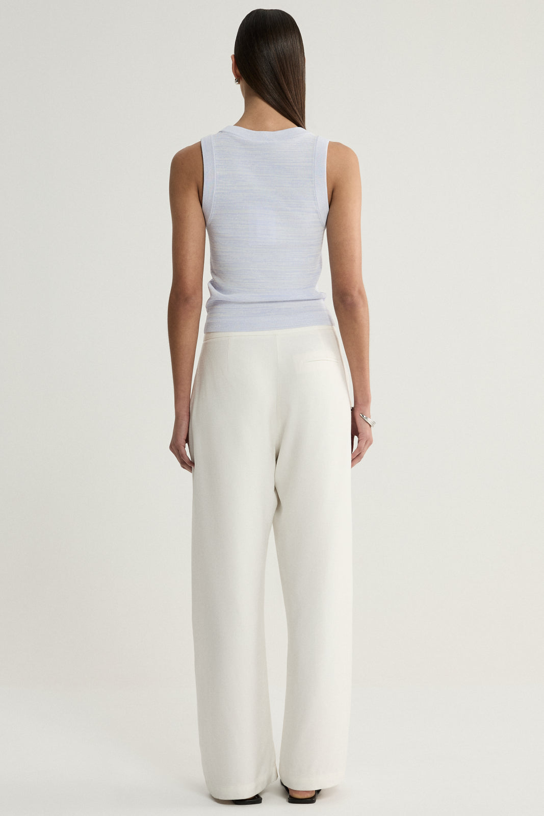 A rear view of a model wearing a pale blue marled sleeveless knit top with a simple back and cream wide-leg trousers, showing the top's fitted shape and hem.