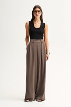 A woman modeling a sleeveless black top and high-waisted, wide-leg taupe-brown trousers against a plain white studio background.
