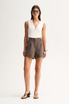 A model wearing taupe tailored mini shorts with front pleats and a white sleeveless V-neck top featuring a short front zip, styled with black strappy sandals in a full-length front view.