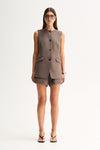 A model wearing taupe tailored mini shorts beneath a matching sleeveless button-front vest, showing the coordinated set with flap-style front pockets and black strappy sandals.