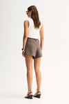 A rear three-quarter view of a model wearing taupe tailored mini shorts with a smooth waistband, belt loops and mock welt pockets, paired with a white sleeveless top and black sandals.