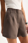A close-up of taupe tailored shorts showing the waistband, belt loops, front pleats, concealed fastening area and angled front pockets.