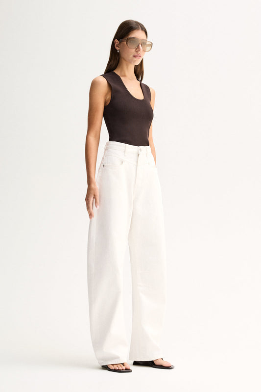 A model in a chocolate-brown sleeveless ribbed top and white wide-leg trousers, photographed from an angled front view that shows the scoop neckline and slim fit through the body.