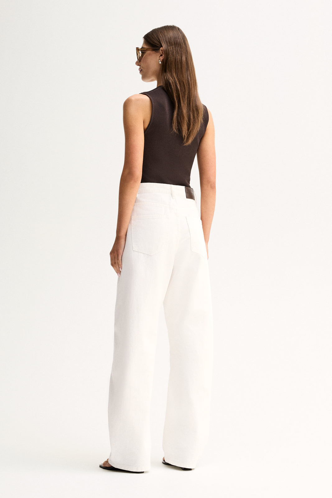 A rear three-quarter view of a model wearing a chocolate-brown sleeveless ribbed top with white wide-leg trousers, showing the back of the top and its regular-length fit.