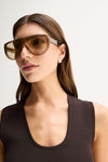 A close-up of a chocolate-brown sleeveless knit top showing its narrow scoop neckline, fine ribbed texture and fitted shoulder line, styled with oversized brown sunglasses.