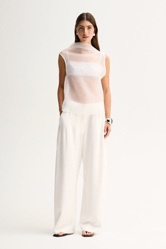 A woman wearing a white sheer mock-neck top with a layered inner slip, styled with wide-leg white trousers in a full-length front view.