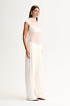 Side view of a woman wearing a white sheer sleeveless top with a mock neck and layered inner slip, paired with wide-leg white trousers.