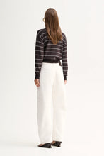 A woman shown from the back wearing a dark striped long-sleeve top and high-waisted white wide-leg trousers, standing against a plain white studio background.