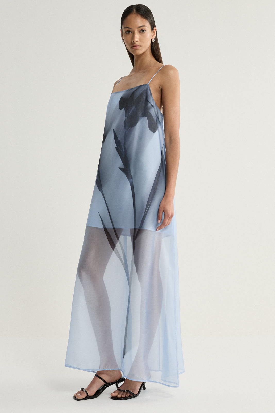 A model wearing a pale blue sheer maxi dress with fine shoulder straps and a large dark floral print, styled with black strappy heels in an angled full-length view.