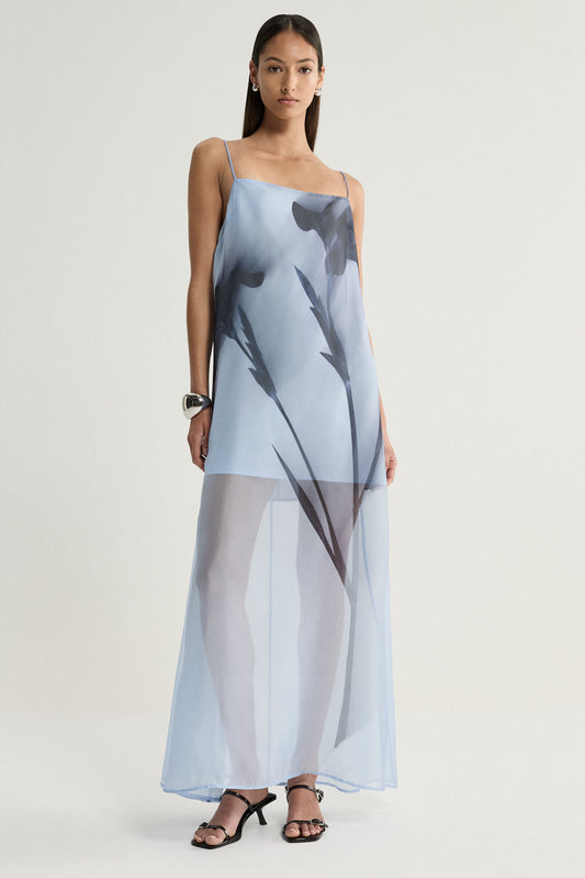 A full-length front view of a pale blue organza maxi dress featuring a square neckline, thin shoulder straps and oversized dark floral shapes, with a sheer flowing skirt and black strappy heels.