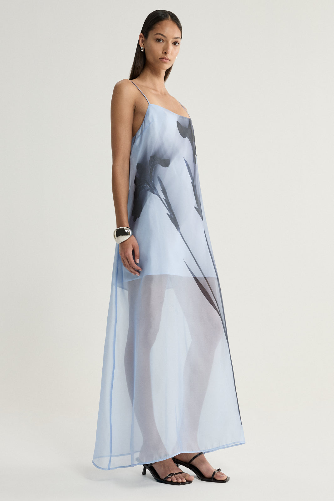 A side view of a model wearing a pale blue sheer maxi dress with a loose straight silhouette, fine shoulder straps and a dark floral print extending down the length of the dress.