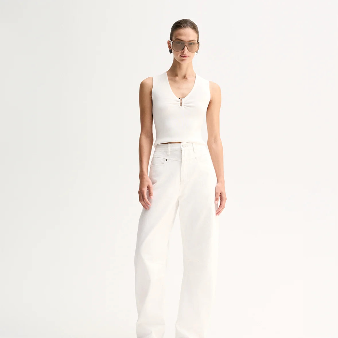 A model wears the white sleeveless knit top with matching wide-leg trousers in a full-length front studio view.
