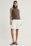 A model wearing a taupe sleeveless top with a high draped cowl neckline, paired with white tailored shorts and black strappy sandals in a full-length front view.