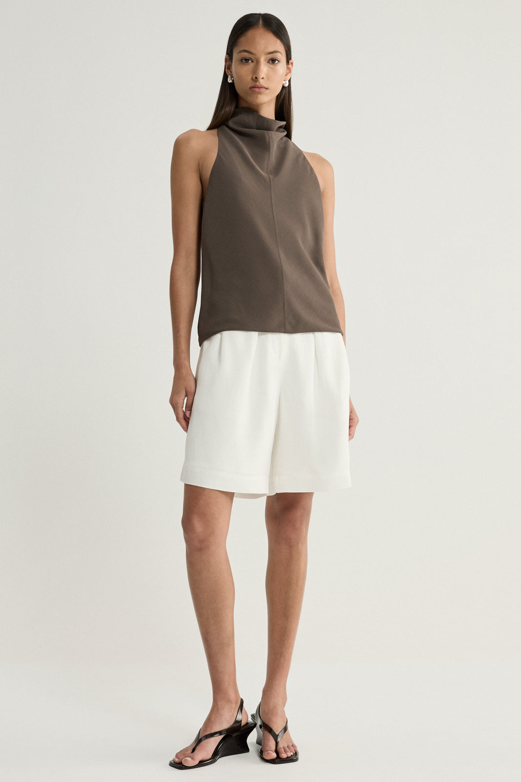 A model wearing a taupe sleeveless top with a high draped cowl neckline, paired with white tailored shorts and black strappy sandals in a full-length front view.