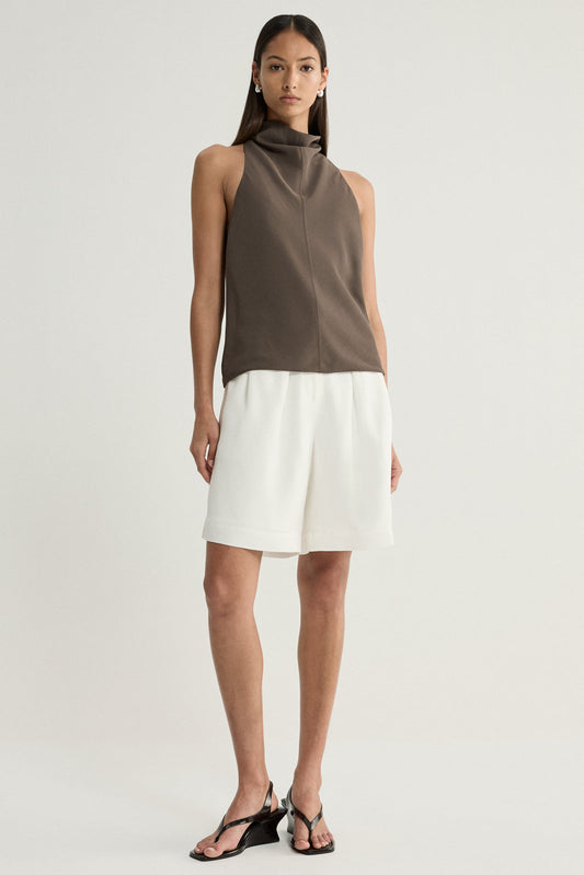 A model wearing a taupe sleeveless top with a high draped cowl neckline, paired with white tailored shorts and black strappy sandals in a full-length front view.