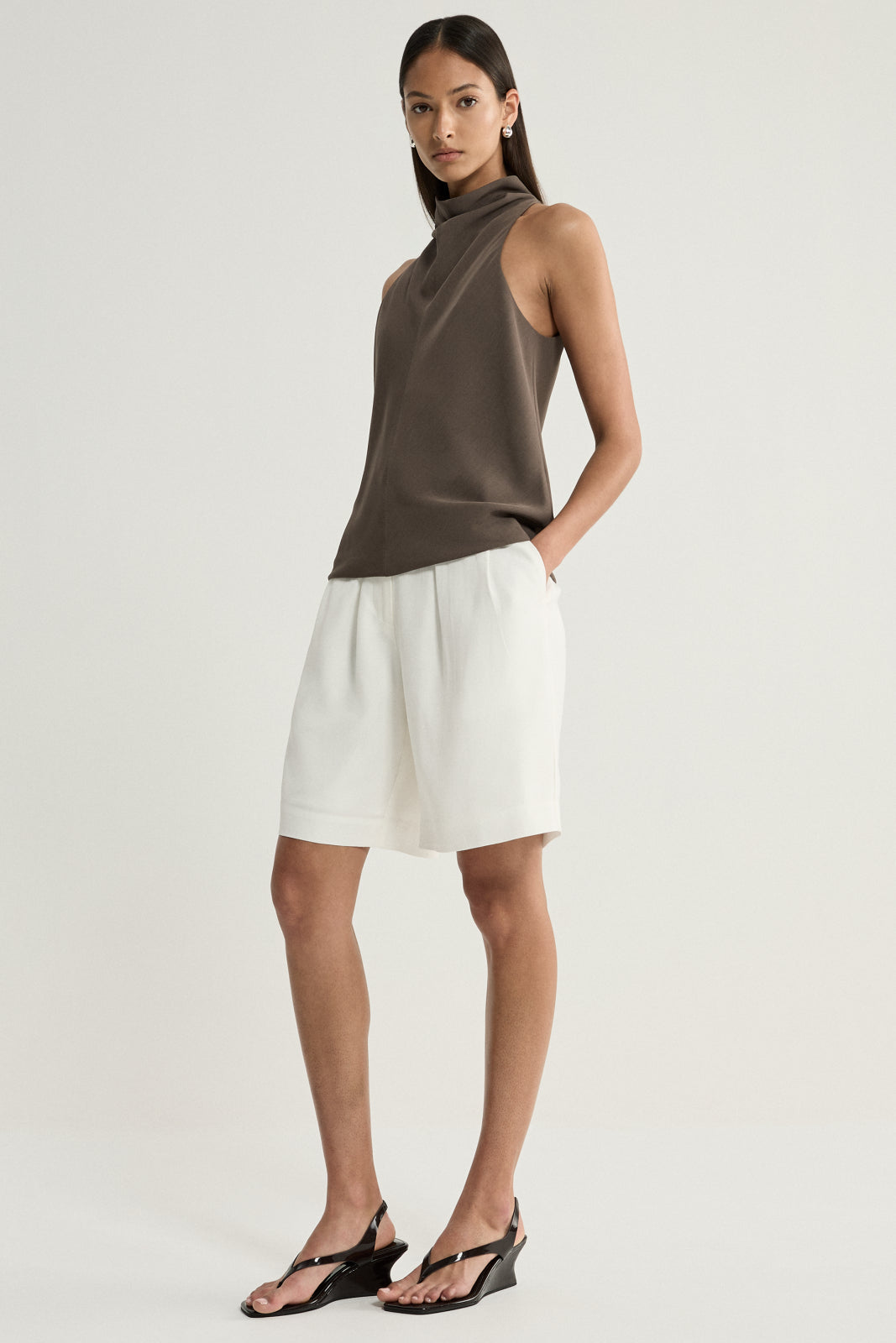 A model wearing a taupe sleeveless top with a high cowl neckline and softly draped front, styled with white tailored shorts and black sandals from an angled front view.