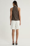 A rear view of a model wearing a taupe sleeveless top with a high neckline and a clean back, paired with white tailored shorts and black heeled sandals.