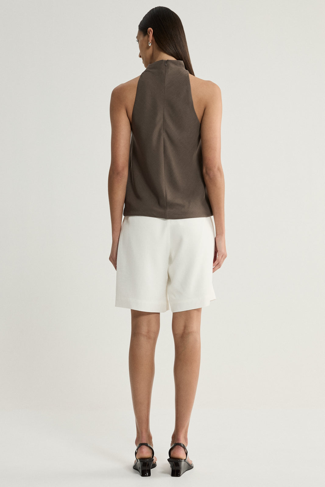 A rear view of a model wearing a taupe sleeveless top with a high neckline and a clean back, paired with white tailored shorts and black heeled sandals.
