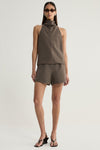 A model wearing a taupe sleeveless top with a sculptural cowl neckline and a straight hem, paired with matching tailored shorts and black strappy sandals.