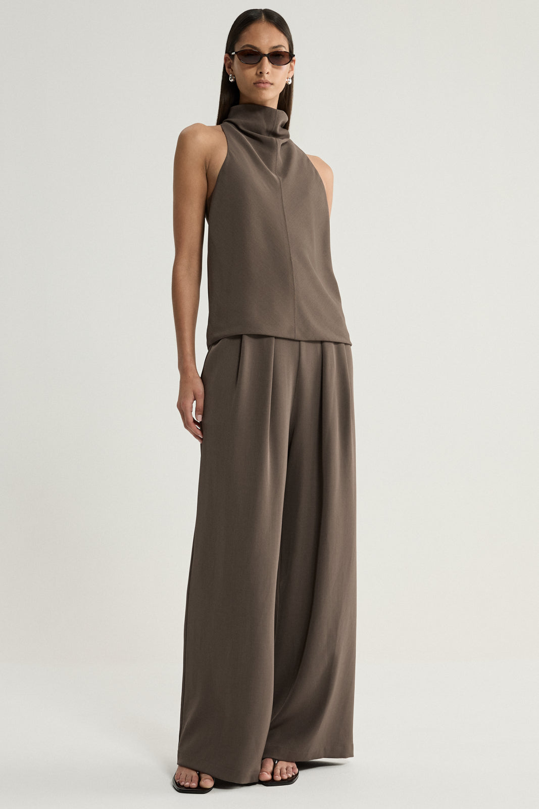A full-length view of a model wearing a taupe sleeveless top with a high draped cowl neckline, styled with matching wide-leg trousers and black sandals.