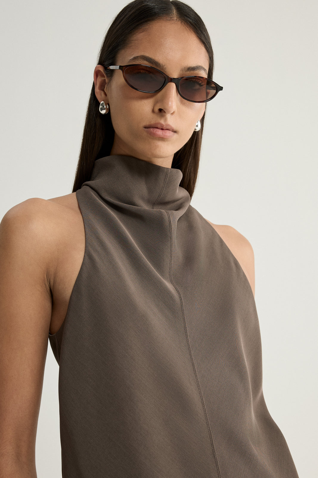 A close-up of a taupe sleeveless top showing the high cowl neckline, softly folded front fabric and clean shoulder line.