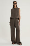 A model wearing a taupe sleeveless vest with a crew neckline, front two-way zip and subtle bubble hem, paired with matching wide-leg trousers and black sandals.
