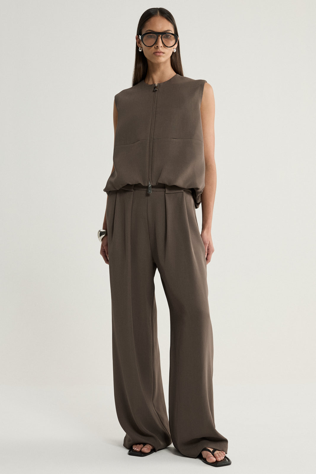 A model wearing a taupe sleeveless vest with a crew neckline, front two-way zip and subtle bubble hem, paired with matching wide-leg trousers and black sandals.