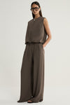 An angled front view of a model wearing a taupe cropped vest with a front zip, rounded crew neckline and softly shaped hem, styled with matching wide-leg trousers.