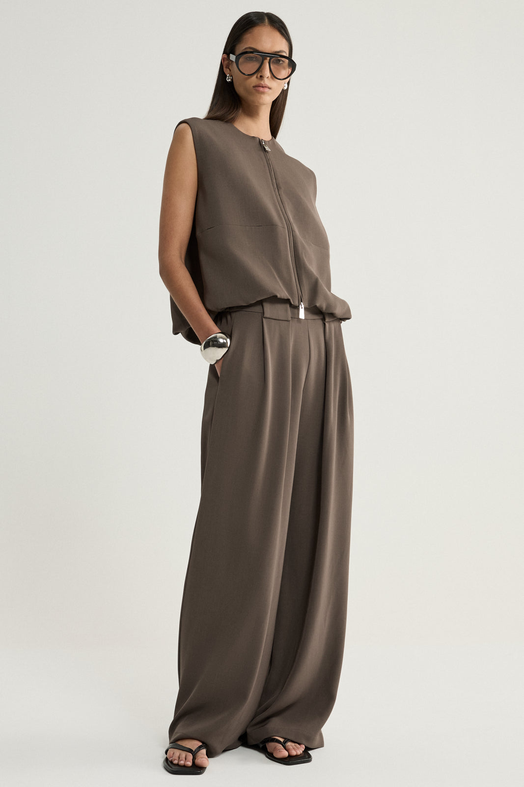 A model wearing a taupe sleeveless zip-front vest with a cropped, softly curved hem and in-seam pockets, shown from a side three-quarter angle with matching wide-leg trousers.
