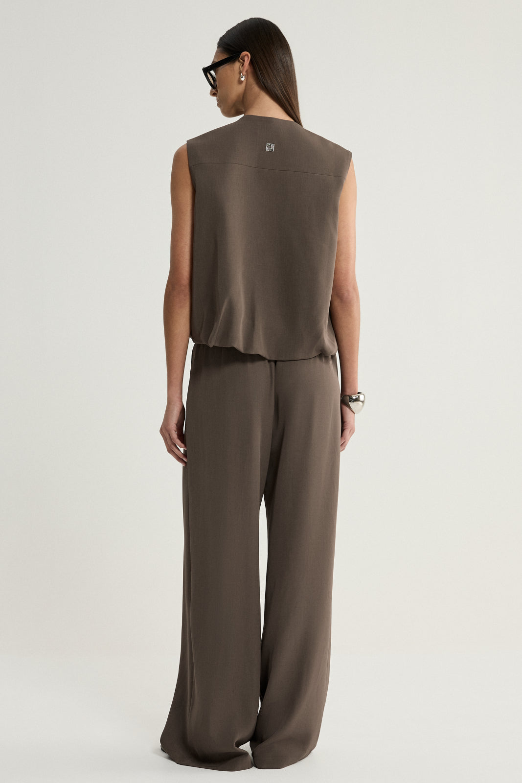 A rear view of a taupe sleeveless vest showing its simple back panel, shoulder seams and cropped hem, styled with matching wide-leg trousers.