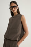 A close-up of a taupe sleeveless vest showing the crew neckline, central two-way zip, horizontal seam across the front and softly structured fabric.