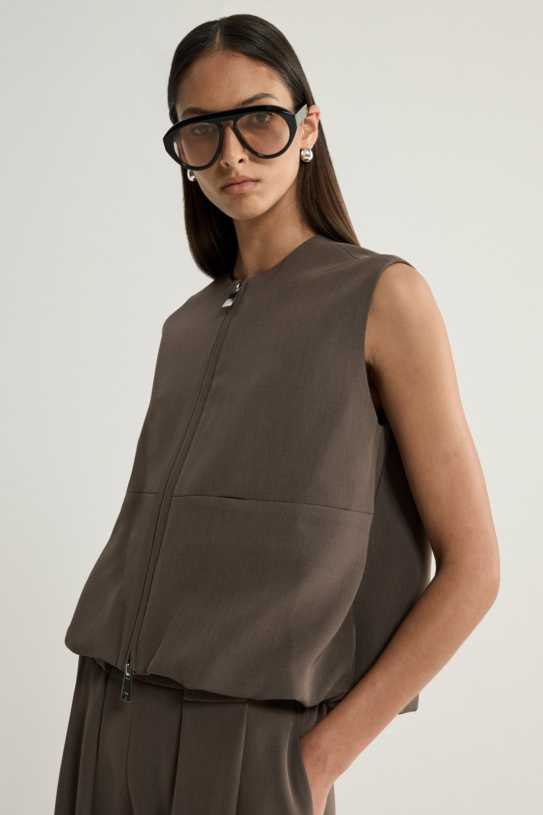 A close-up of a taupe sleeveless vest showing the crew neckline, central two-way zip, horizontal seam across the front and softly structured fabric.