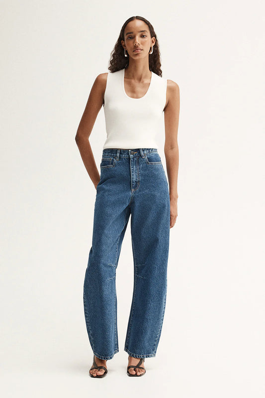 Model wearing blue high-rise barrel-leg jeans with a fitted white tank top and black sandals, showing the curved leg silhouette and full-length fit, front view