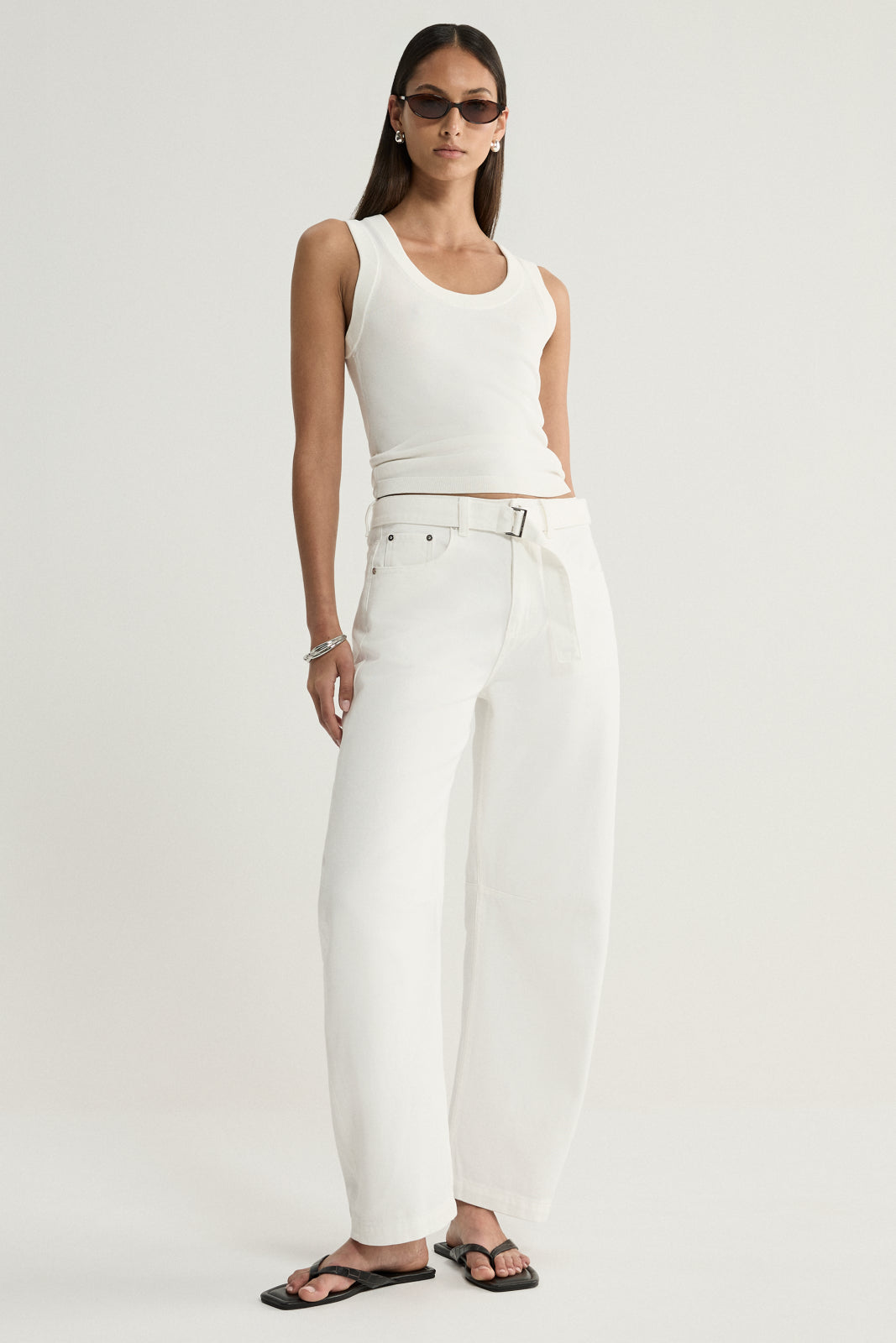 A model wearing ivory wide-leg denim pants with a removable tie belt, styled with a white sleeveless top, dark sunglasses and black flat sandals in a full-length front view.