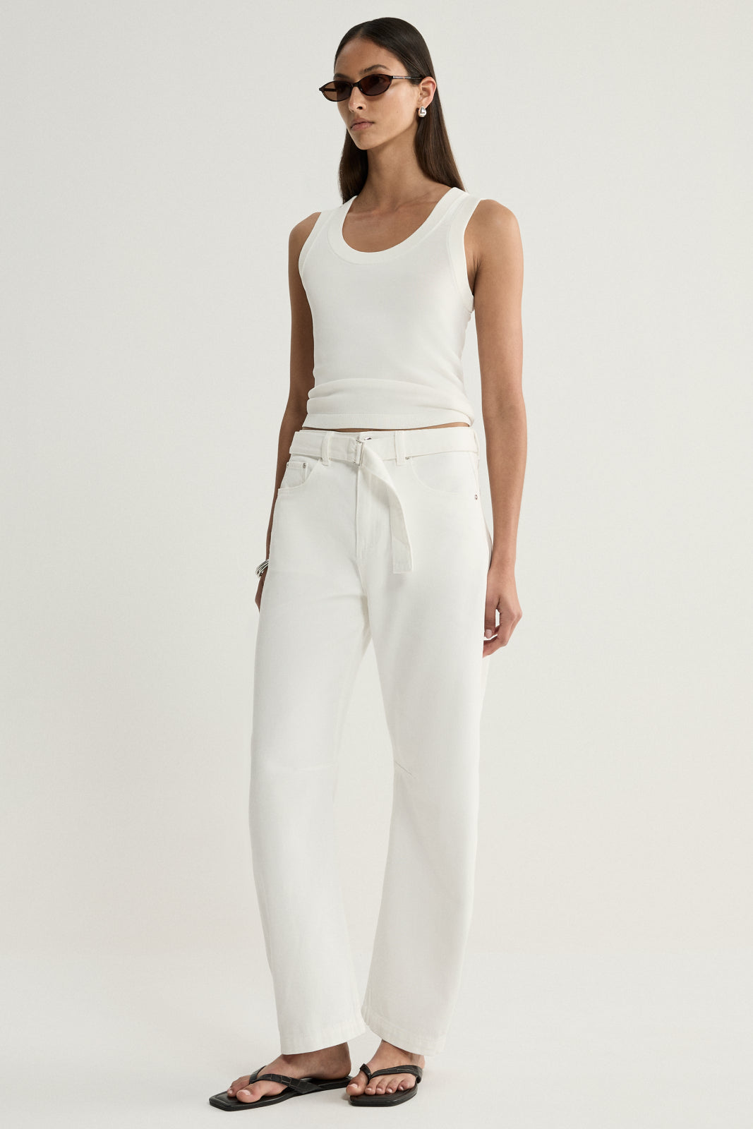 A model wearing ivory wide-leg denim pants with a tied waist belt and front pockets, paired with a white sleeveless top and black sandals in an angled front pose.