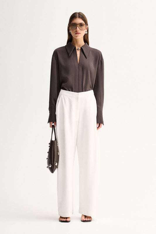 Front three-quarter view of a woman wearing a slate collared shirt with long sleeves and buttoned cuffs, paired with white trousers.