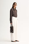 Side view of a woman wearing a slate long-sleeve shirt with a fluid drape and white wide-leg trousers.
