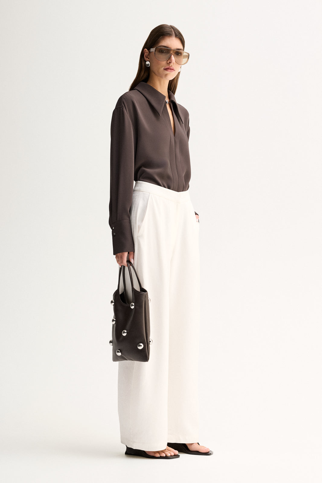 Side view of a woman wearing a slate long-sleeve shirt with a fluid drape and white wide-leg trousers.