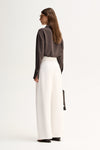 Back three-quarter view of a woman wearing a slate long-sleeve shirt with a relaxed drape and white trousers.