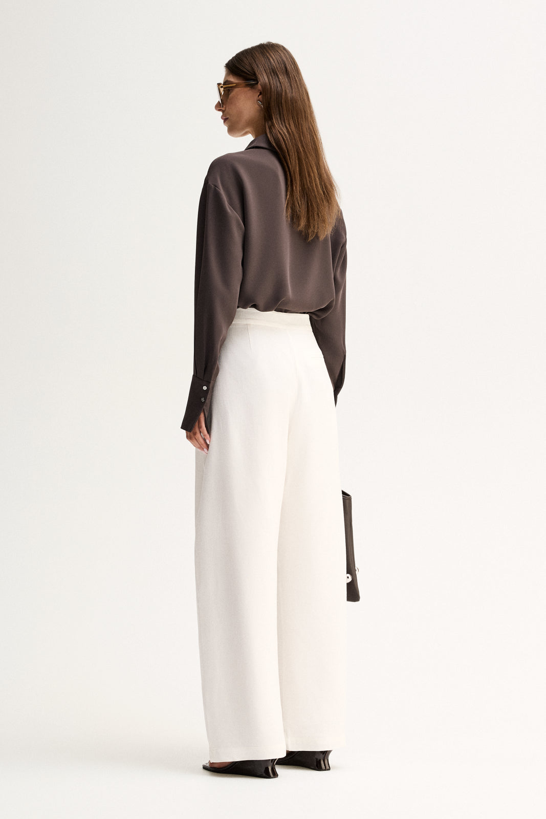 Back three-quarter view of a woman wearing a slate long-sleeve shirt with a relaxed drape and white trousers.