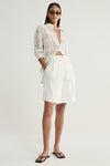 A model wearing a white semi-sheer check shirt with a pointed collar, front buttons and rolled sleeves, styled open at the neckline with ivory tailored shorts and black flat sandals.