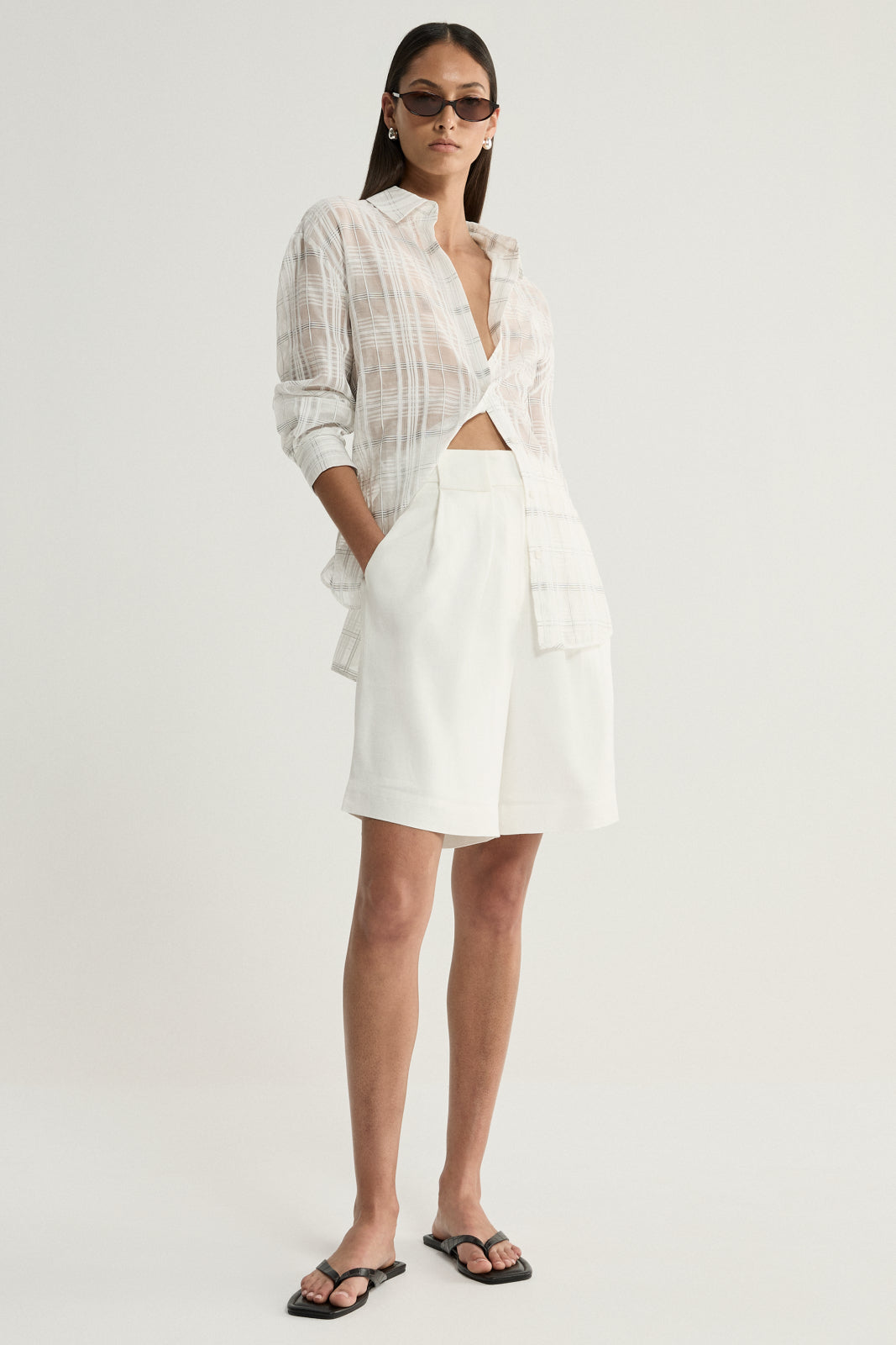 A model wearing a white semi-sheer check shirt with a pointed collar, front buttons and rolled sleeves, styled open at the neckline with ivory tailored shorts and black flat sandals.