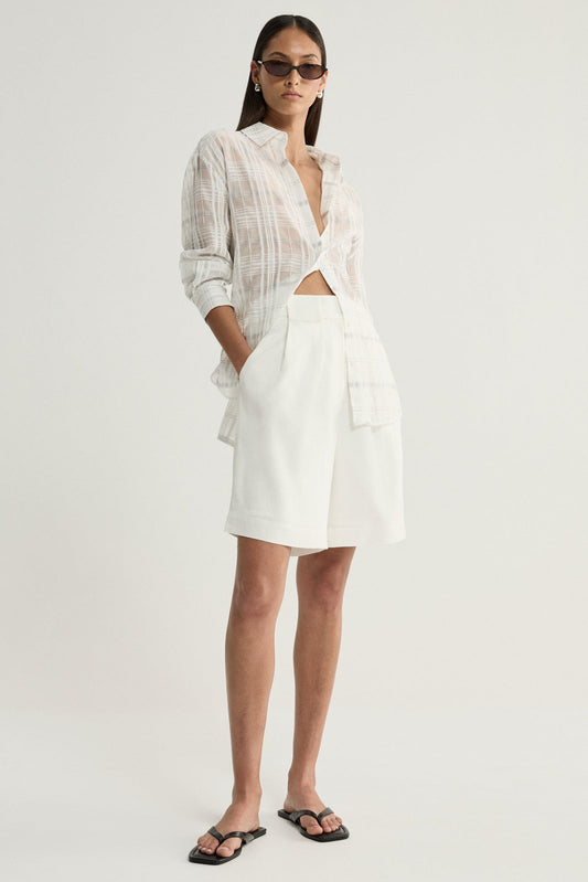 A model wearing a white semi-sheer check shirt with a pointed collar, front buttons and rolled sleeves, styled open at the neckline with ivory tailored shorts and black flat sandals.