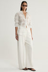 A model wearing a white sheer check shirt with neutral vertical and horizontal lines, a collared neckline and long sleeves, paired with ivory wide-leg trousers and black flat sandals in a full-length front view.