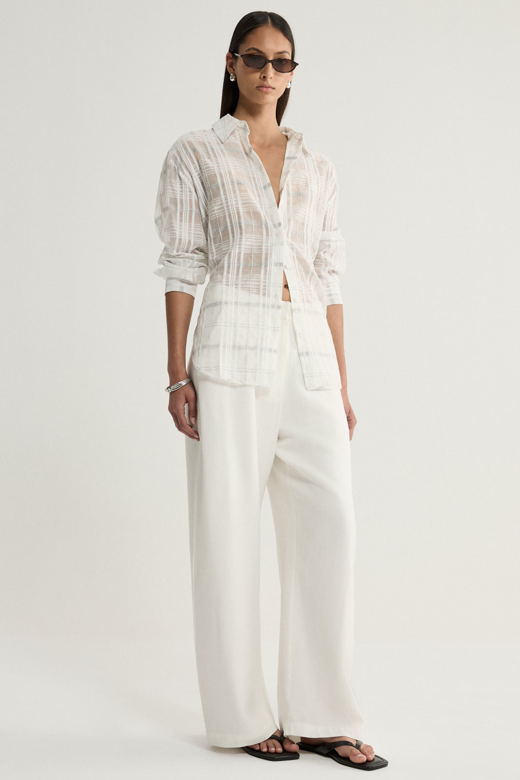 A model wearing a white sheer check shirt with neutral vertical and horizontal lines, a collared neckline and long sleeves, paired with ivory wide-leg trousers and black flat sandals in a full-length front view.