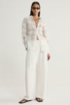 A model wearing a white semi-sheer checked button-up shirt with an open collar and relaxed sleeves, styled with ivory wide-leg trousers and black sandals in a straight-on studio pose.